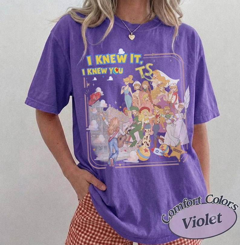 May include: A purple short-sleeve t-shirt featuring a graphic print of various animated characters in a collage style. The text "I KNEW IT, I KNEW YOU TS" is printed in yellow and blue above the characters. The design includes a sheriff star.