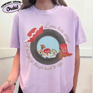 May include: A light purple t-shirt featuring a graphic of a black tire framing a scene with three cartoon characters. A red cowboy hat sits on the tire, and a red cowboy boot is on the right. Text reads: Love has ways of bringing things back to life.