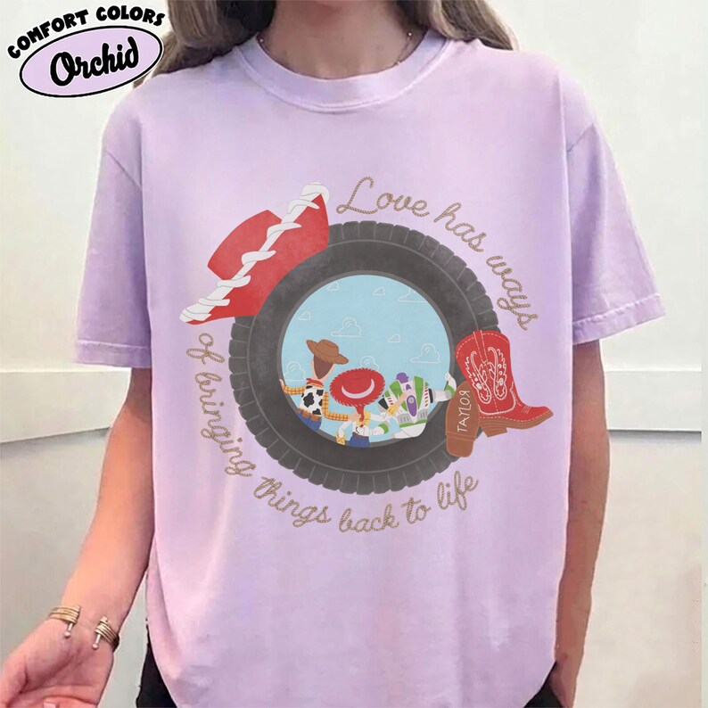 May include: A light purple t-shirt featuring a graphic of a black tire framing a scene with three cartoon characters. A red cowboy hat sits on the tire, and a red cowboy boot is on the right. Text reads: Love has ways of bringing things back to life.
