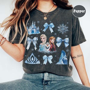 May include: Dark grey t-shirt with a Frozen-themed design, including Elsa, Anna, Olaf, blue bows, snowflakes, and a castle. The tee has a vintage wash and a crew neck.
