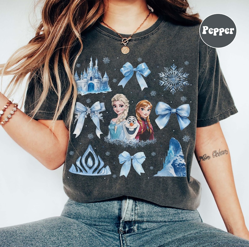 May include: Dark grey t-shirt with a Frozen-themed design, including Elsa, Anna, Olaf, blue bows, snowflakes, and a castle. The tee has a vintage wash and a crew neck.