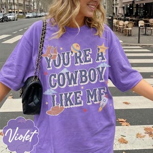 May include: A purple short-sleeve t-shirt featuring the text "YOU'RE A COWBOY LIKE ME" in a distressed font. The graphic includes illustrations of cowboy boots, a ball, a sheriff star, clouds, a cowboy hat, and a rocket ship with small stars.