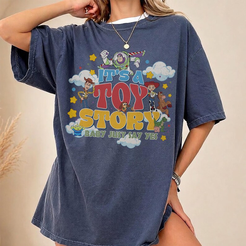 May include: A dark blue short-sleeve t-shirt featuring a graphic print of Toy Story characters including Buzz Lightyear, Woody, and Jessie. The text reads IT'S A TOY STORY BABY JUST SAY YES, surrounded by yellow stars and white clouds.