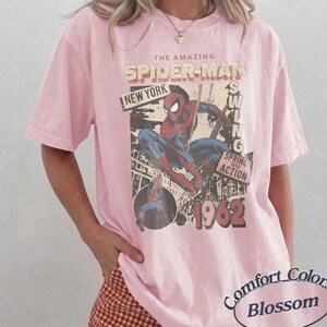 May include: A light pink short-sleeve t-shirt featuring a graphic of Spider-Man in a dynamic pose. The print includes the text THE AMAZING SPIDER-MAN, NEW YORK, SWING, SPRING INTO ACTION, and 1962. The shirt has a crew neckline and a relaxed fit.