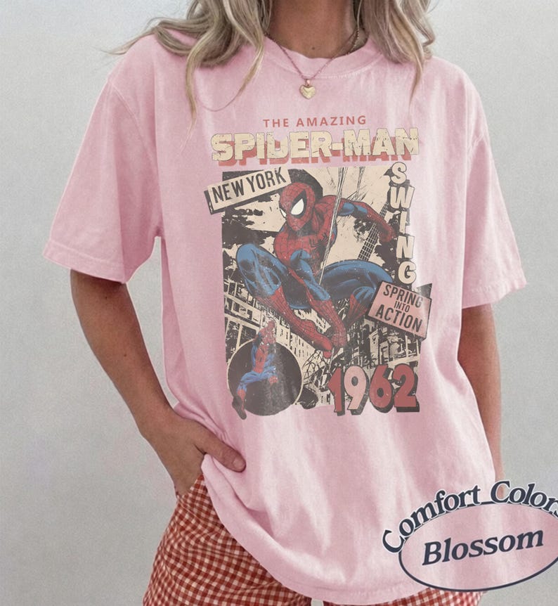 May include: A light pink short-sleeve t-shirt featuring a graphic of Spider-Man in a dynamic pose. The print includes the text THE AMAZING SPIDER-MAN, NEW YORK, SWING, SPRING INTO ACTION, and 1962. The shirt has a crew neckline and a relaxed fit.