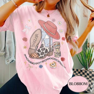 Puede incluir: Una camiseta rosa claro con un estampado gr&aacute;fico de una bota de vaquero con patr&oacute;n de vaca, un sombrero rojo, bloques de letras con la T, la S y el n&uacute;mero 13, un lazo y una cadena de cuentas con el texto I KNEW IT I KNEW YOU.