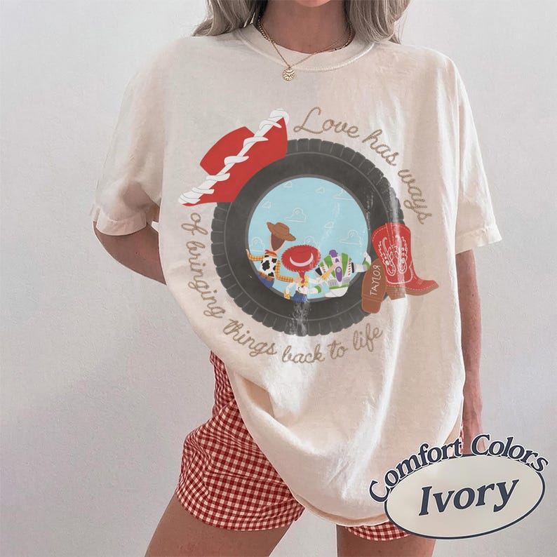 May include: An ivory t-shirt featuring a graphic of a black tire framing a scene with cartoon characters, a red cowboy hat, and a red boot. The text reads, Love has ways of bringing things back to life. A small label in the corner says Comfort Colors Ivory.