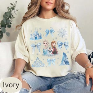 May include: Ivory t-shirt with a colourful design featuring a castle, snowflakes, bows, and characters from a popular animated movie. The design includes Elsa, Anna, and Olaf in shades of blue and white.