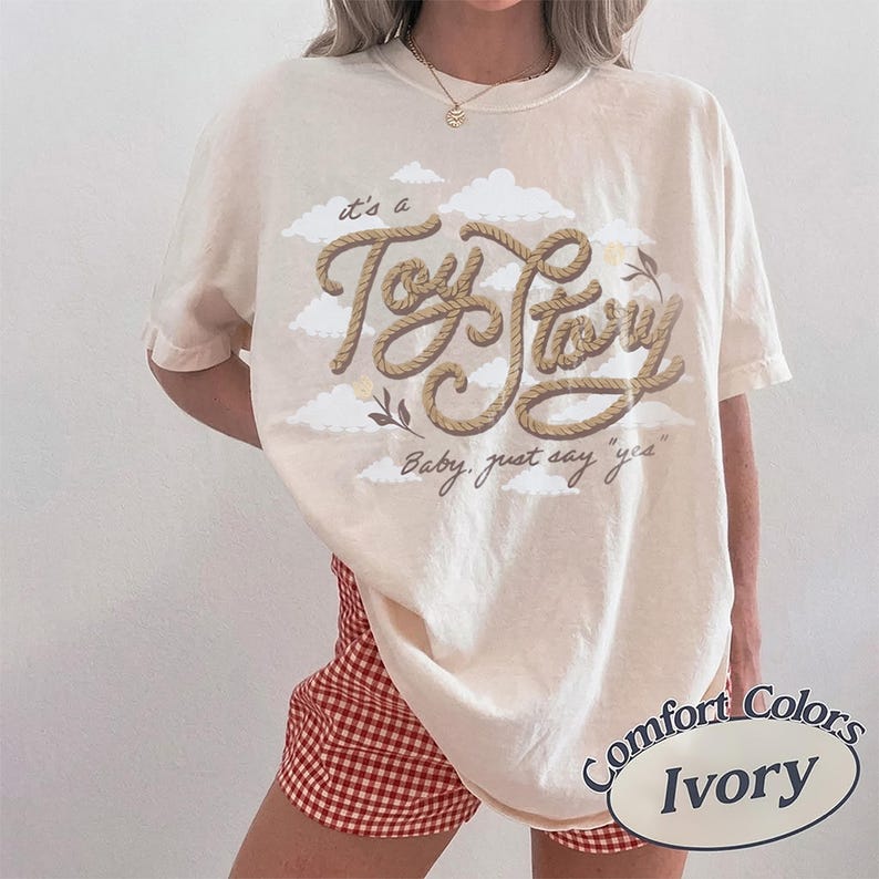 Pu&ograve; includere: Una t-shirt a maniche corte color avorio con una grafica di nuvole bianche e il testo "it's a Toy Story" scritto in un carattere marrone stile corda, con il testo "Baby, just say "yes"" stampato sotto in un carattere corsivo marrone pi&ugrave; piccolo.