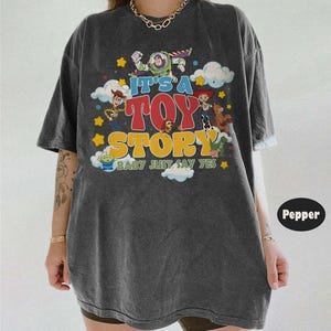 May include: A charcoal gray short-sleeve t-shirt featuring a colorful graphic print of Toy Story characters including Buzz Lightyear, Woody, Jessie, and others. The text reads IT'S A TOY STORY BABY JUST SAY YES, surrounded by clouds and stars.