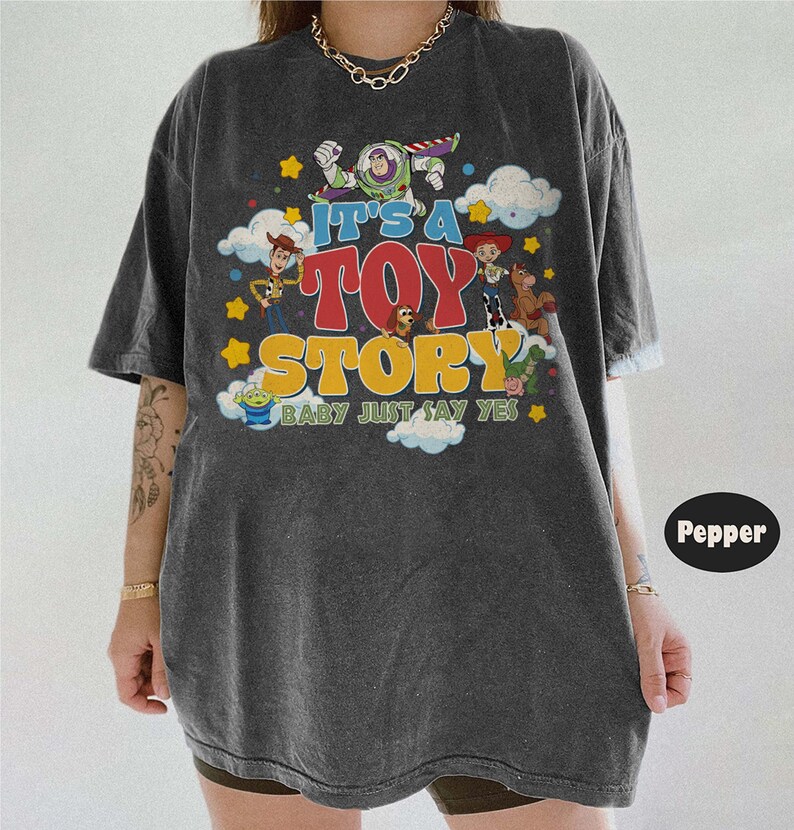 May include: A charcoal gray short-sleeve t-shirt featuring a colorful graphic print of Toy Story characters including Buzz Lightyear, Woody, Jessie, and others. The text reads IT'S A TOY STORY BABY JUST SAY YES, surrounded by clouds and stars.