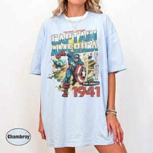 Puede incluir: Una camiseta azul claro con un gr&aacute;fico de estilo vintage que muestra al Capit&aacute;n Am&eacute;rica en una pose din&aacute;mica con un escudo. El texto dice THE FIRST AVENGER CAPTAIN AMERICA FIGHT FOR LIBERTY 1941. El dise&ntilde;o incluye detalles desgastados.