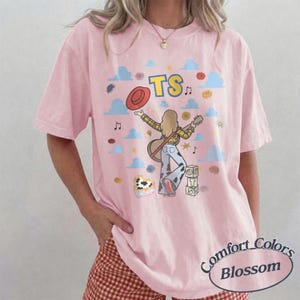 Op de afbeelding: Een lichtroze T-shirt met korte mouwen en een afbeelding van een persoon met lang haar die gitaar speelt, gekleed in een geruit overhemd en een broek met koeienprint. Het ontwerp bevat een rode hoed, blauwe wolken en de tekst TS.