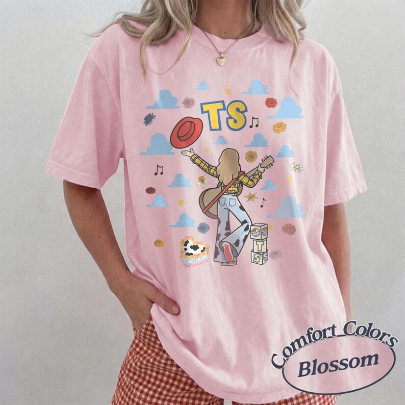 Op de afbeelding: Een lichtroze T-shirt met korte mouwen en een afbeelding van een persoon met lang haar die gitaar speelt, gekleed in een geruit overhemd en een broek met koeienprint. Het ontwerp bevat een rode hoed, blauwe wolken en de tekst TS.