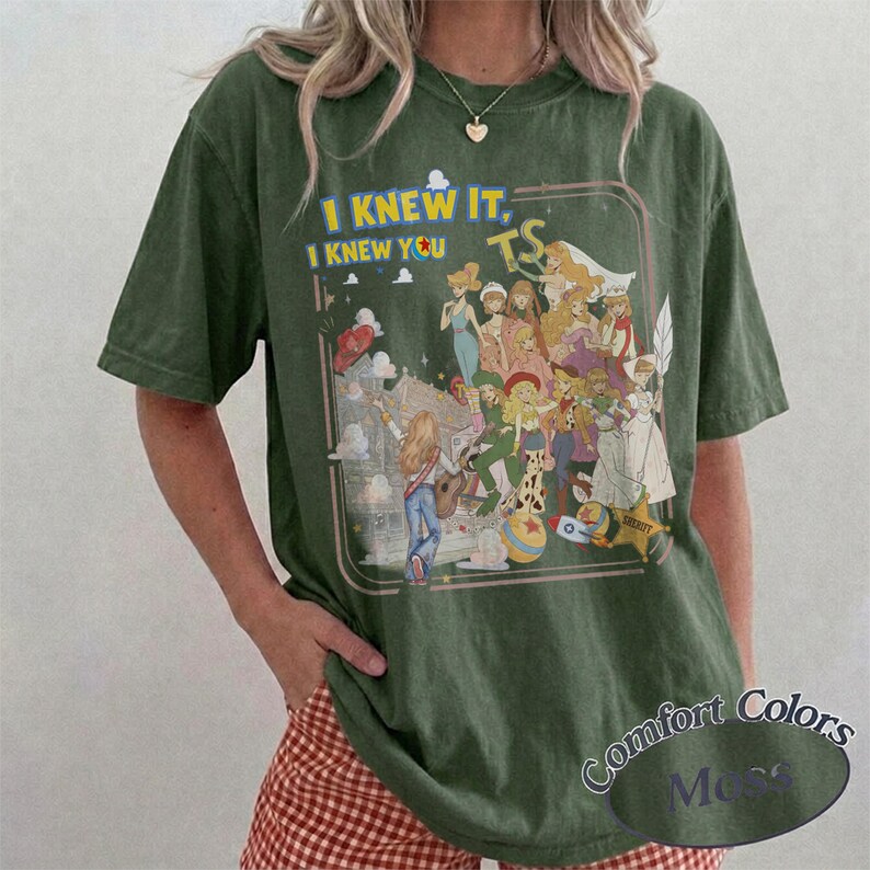 May include: A dark olive green short-sleeve t-shirt featuring a graphic print of various animated characters. The text "I KNEW IT, I KNEW YOU" appears in yellow and blue above the characters, with "TS" in yellow. The design includes a star and clouds.