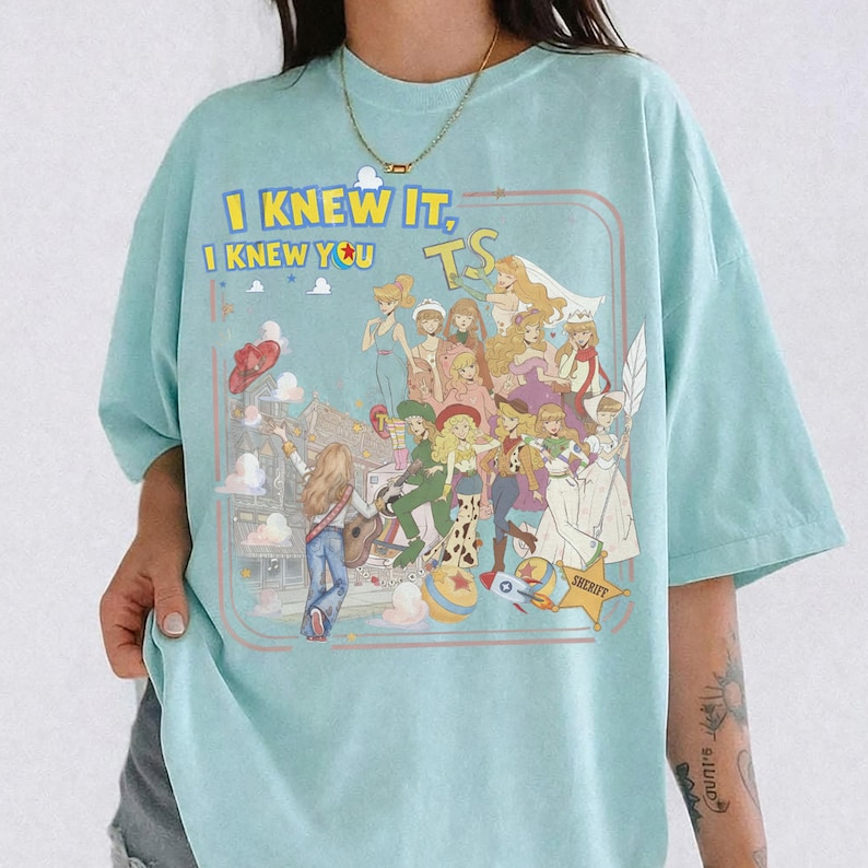 May include: A light blue t-shirt featuring a graphic print of various animated characters in a collage style. The text "I KNEW IT, I KNEW YOU" is printed in yellow above the characters, with a small "TS" logo and a yellow sheriff star badge.