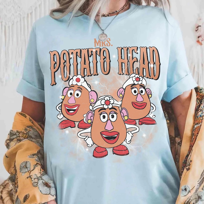 Op de afbeelding: Lichtblauw T-shirt met een afbeelding van drie Mrs. Potato Head-personages en de tekst "MRS. POTATO HEAD" in een retro lettertype. Het ontwerp bevat de gezichten, hoeden en rode schoenen van de personages.