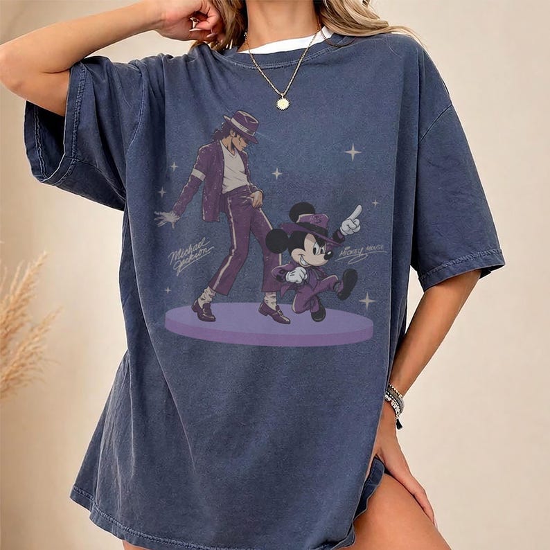 Pode incluir: Uma t-shirt azul-escura oversized com uma estampa gr&aacute;fica de Michael Jackson e Mickey Mouse, ambos vestindo ternos e chap&eacute;us fedora roxos. Eles dan&ccedil;am em uma plataforma circular roxa cercada por pequenas estrelas brancas e texto em cursivo.
