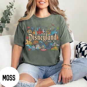 May include: Moss green t-shirt featuring a colorful Disneyland Resort graphic. The design includes the Disneyland logo and various park icons. The shirt is a short-sleeved, crew neck style.