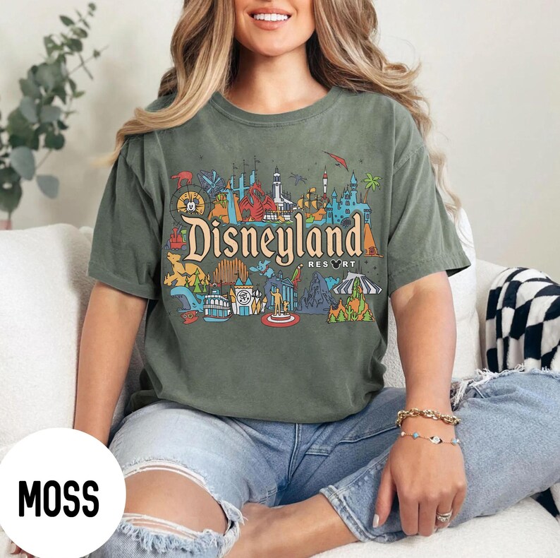 May include: Moss green t-shirt featuring a colorful Disneyland Resort graphic. The design includes the Disneyland logo and various park icons. The shirt is a short-sleeved, crew neck style.