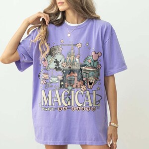 Puede incluir: Una camiseta de color lila con un dise&ntilde;o gr&aacute;fico que dice "The Most Magical Place on Earth". El dise&ntilde;o incluye ilustraciones de un castillo, tazas de t&eacute; y otros elementos de parques tem&aacute;ticos. La camiseta tiene mangas cortas y cuello redondo.