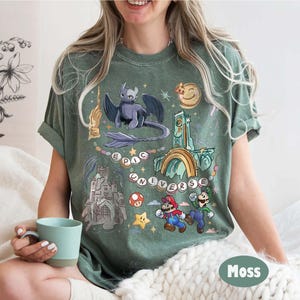 May include: A moss green t-shirt featuring a colorful graphic design. The design includes a dragon, a castle, and the words "EPIC UNIVERSE". The shirt also features cartoon characters Mario and Luigi.