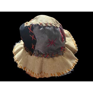 May include: A patchwork bucket hat constructed from beige burlap, black fabric, and grey suede-like material. The hat features decorative cross-stitch embroidery in red and orange thread along the seams and edges, with a wide, frayed-edge brim.