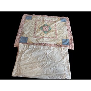 May include: A cream-colored quilt with a floral and blue patchwork design. The quilt features a central diamond pattern and blue square accents. The edges are trimmed with a floral pattern. A matching cream-colored pillowcase is visible.