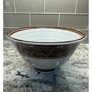 May include: A ceramic bowl with a wide, flared rim and a tapered base. The interior is a smooth, off-white glaze. The exterior features a dark brown band around the upper section with a carved, repeating leaf-like pattern, set against a light cream base.
