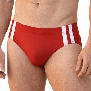 May include: A pair of red swim briefs featuring two vertical white stripes on each side. The garment has a solid red base color with a clean, minimalist design and a standard cut, providing a classic look for swimming and water-based activities.