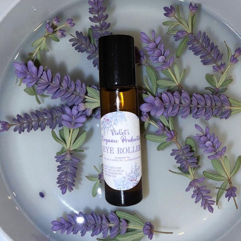 Pu&ograve; includere: Un flacone roll-on per occhi in vetro marrone con tappo nero, etichettato "Violet's Organic Products EYE ROLLER". Il flacone &egrave; circondato da rametti di fiori di lavanda viola e foglie verdi, disposti su una superficie azzurra.