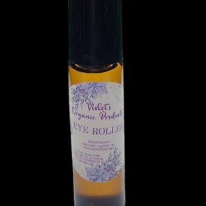 Pu&ograve; includere: Un flacone roll-on per occhi in vetro marrone con tappo nero. L'etichetta recita "Violet's Organic Products EYE ROLLER" con un design floreale. Gli ingredienti includono olio di ricino biologico e olio di incenso.