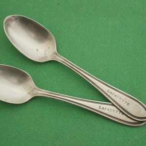 Puede incluir: Dos cucharas de plata con la palabra "LAFAYETTE" grabada en el mango, sobre una superficie verde. Las cucharas tienen un cuenco ovalado y una línea decorativa a lo largo del mango.