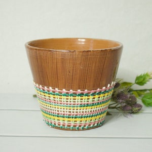 Puede incluir: Una maceta de cerámica marrón con un diseño tejido de colores alrededor de la parte superior. La maceta está sobre una superficie blanca.