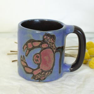 May include: A blue ceramic mug with a crab design. The mug has a black handle and interior, and a pink crab illustration on the front. The mug is on a white surface with yellow flowers.