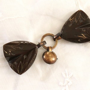 May include: A brown, carved, wooden, triangular-shaped, decorative clasp with a gold-colored metal ring and a bell charm.