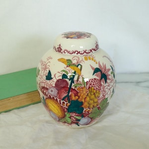 May include: A white ceramic jar with a lid, adorned with a colorful fruit and floral design. The jar features a round shape and is decorated with red, yellow, green, and purple hues. A green book is visible in the background.