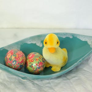 Könnte beinhalten: Eine tealblaue, keramik-gefertigte, blattförmige Servierplatte mit weißem Überzug, die zwei bunte, gemusterte Ostereier und eine gelbe Gummiente enthält.