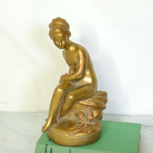 May include: A gold-coloured statue of a seated figure, possibly a classical nymph, resting on a textured base. The statue is placed on a green book. The figure is detailed with flowing hair and a serene expression. The statue is approximately 20 cm tall.