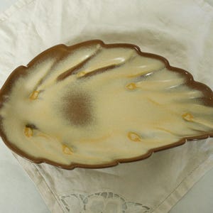 May include: A leaf-shaped ceramic serving dish with a brown rim and a cream-coloured interior. The dish has a textured surface and is resting on a white cloth. The dish is approximately 30 cm long.