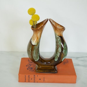 Peut inclure: Un vase en céramique en forme de U, avec un glaçage marron, vert et blanc. Le vase contient des fleurs jaunes sphériques et repose sur un livre orange vintage. Le vase mesure environ 20 cm de haut.