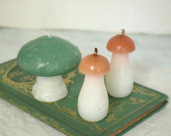 May include: Three mushroom-shaped candles in varying sizes and colours. One is green with a white stem, and two are white with pink and brown caps. The candles are placed on a green book with gold lettering.