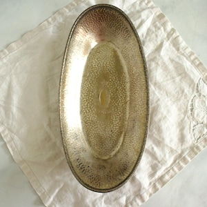 May include: An oval, silver-toned metal serving tray with a textured interior and a decorative rim. The tray rests on a cream-coloured linen napkin with a floral embroidered corner. The tray is empty.