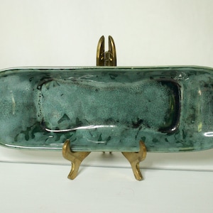 Peut inclure: Un plat de service en céramique vert avec un glaçage moucheté. Le plat est de forme ovale et présente une légère courbe au centre. Il est posé sur un support en métal doré.