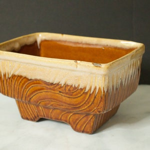 Puede incluir: Un macetero rectangular de cerámica con un esmalte marrón y blanco. El macetero tiene un patrón ondulado de veta de madera y una base ligeramente elevada.