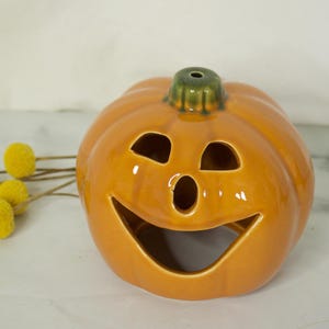 Puede incluir: Un portavelas de cerámica con forma de calabaza sonriente. La calabaza naranja tiene ojos y boca recortados, y un tallo verde. Flores amarillas de pompón en el fondo. Una decoración festiva para Halloween.