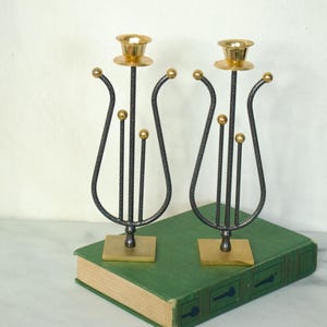May include: A pair of black metal candle holders with gold accents. The candle holders are shaped like a lyre and have a minimalist design.