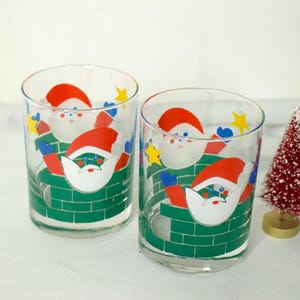 May include: Two clear glass tumblers with a Christmas theme. Each glass features a green brick chimney with Santa Claus climbing down it. The Santa Claus figures are wearing red hats, white beards, and blue and red clothing.