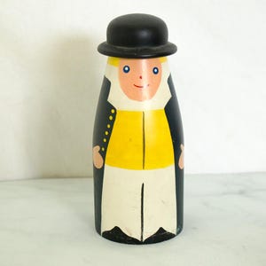 May include: A wooden figurine of a person wearing a black top hat, a black jacket, a yellow vest, and white pants. The figurine is painted with a smiling face and blue eyes.
