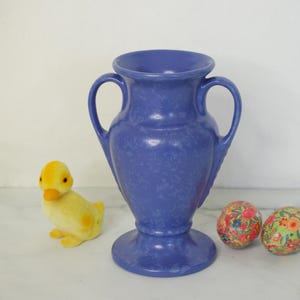 May include: A blue vase with two handles, a yellow duck figurine, and two decorative eggs with floral patterns. The vase has a speckled texture and a wide base. The duck is plush and the eggs are round.
